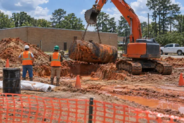 Rusty underground fuel tank lifted from a red clay excavation at a small-town commercial lot on a sunny day