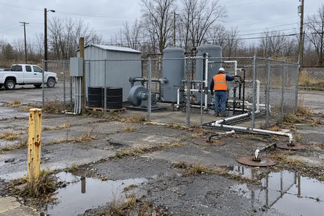 Remediation treatment system inside a chain-link enclosure on a former gas station lot, with a blower, piping and treatment vessels