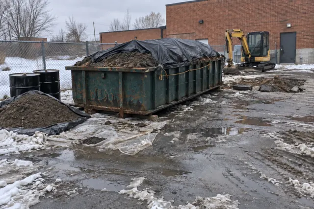 Roll-off container of excavated soil covered with a black tarp at the back of a commercial lot